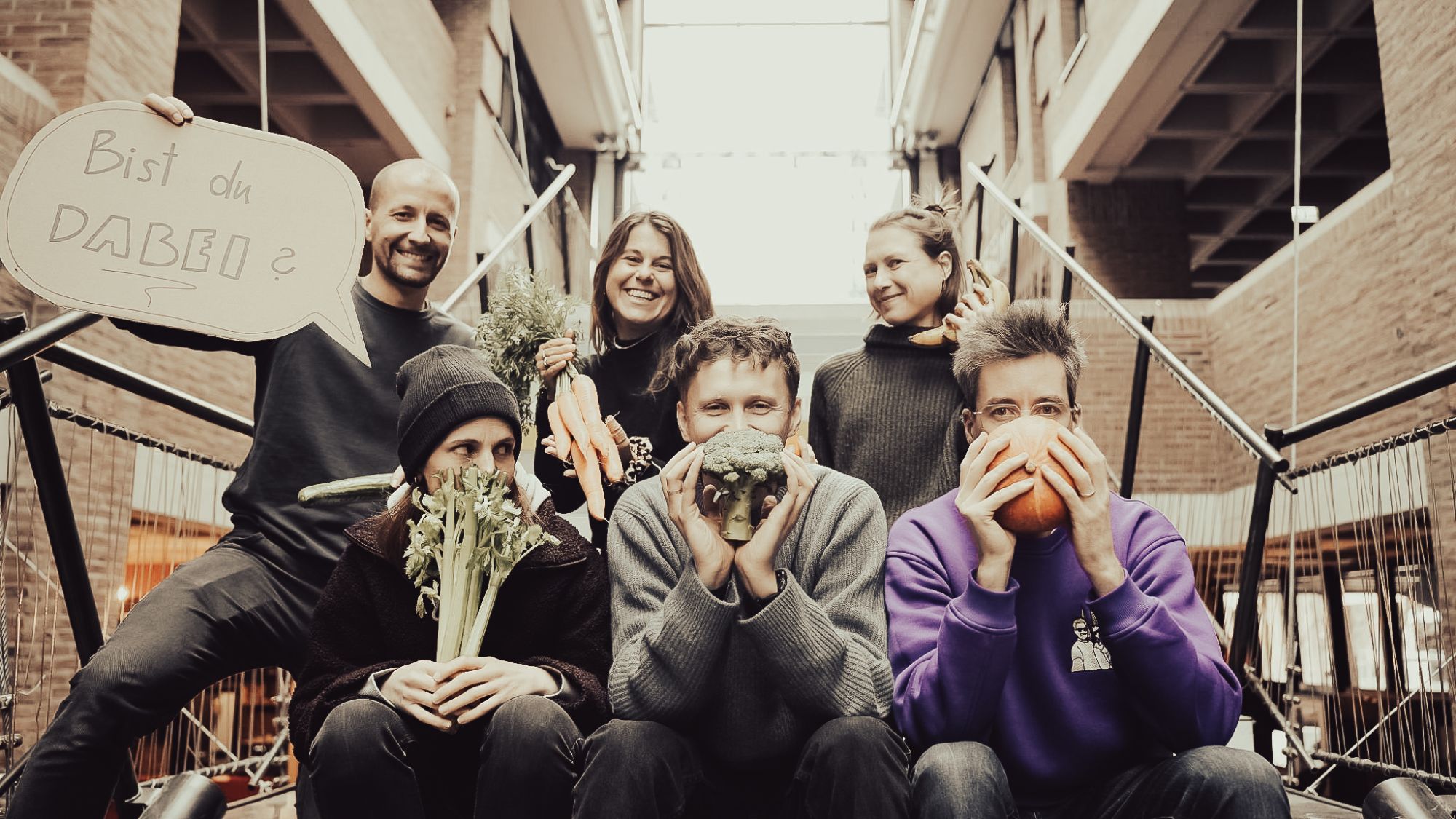 Das Kollektiv hinter den Formaten: Gruppenfoto, alle halten sich Gemüse vors Gesicht, ein Schild fragt „Bist du dabei?“